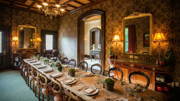 Een formele eetzaal met een lange tafel gedekt voor een maaltijd, omringd door stoelen en versierd met potplanten.