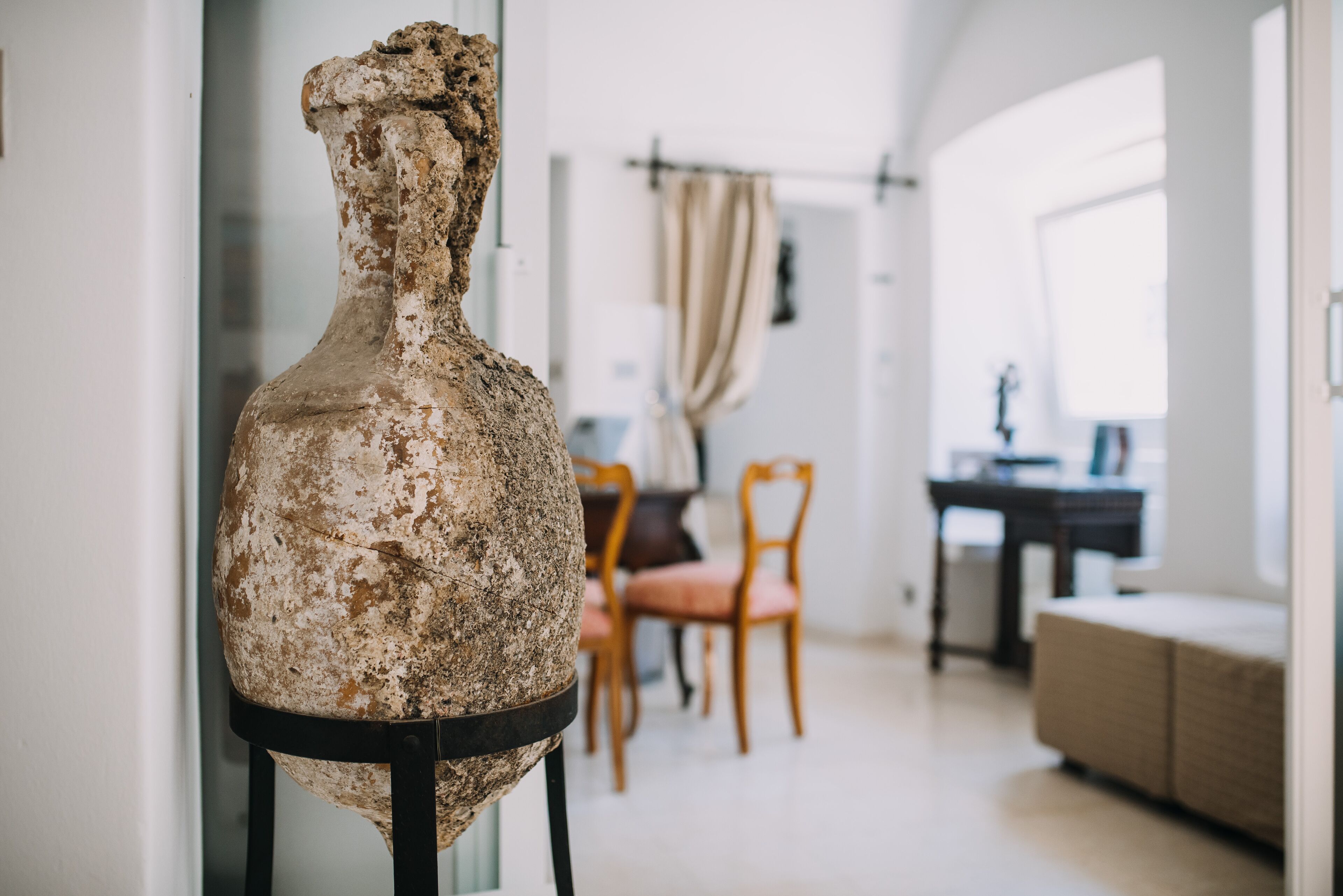 Una habitación con un jarrón rústico y desgastado sobre un pedestal, un comedor con sillas y una mesa, y una ventana con cortinas.