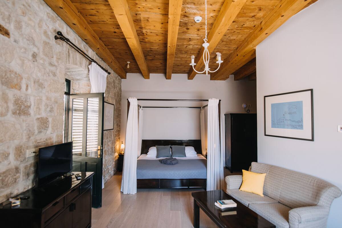 Een slaapkamer met een stenen muur, houten balken in het plafond, een hemelbed, een televisie, een bank en een ingelijst schilderij aan de muur.
