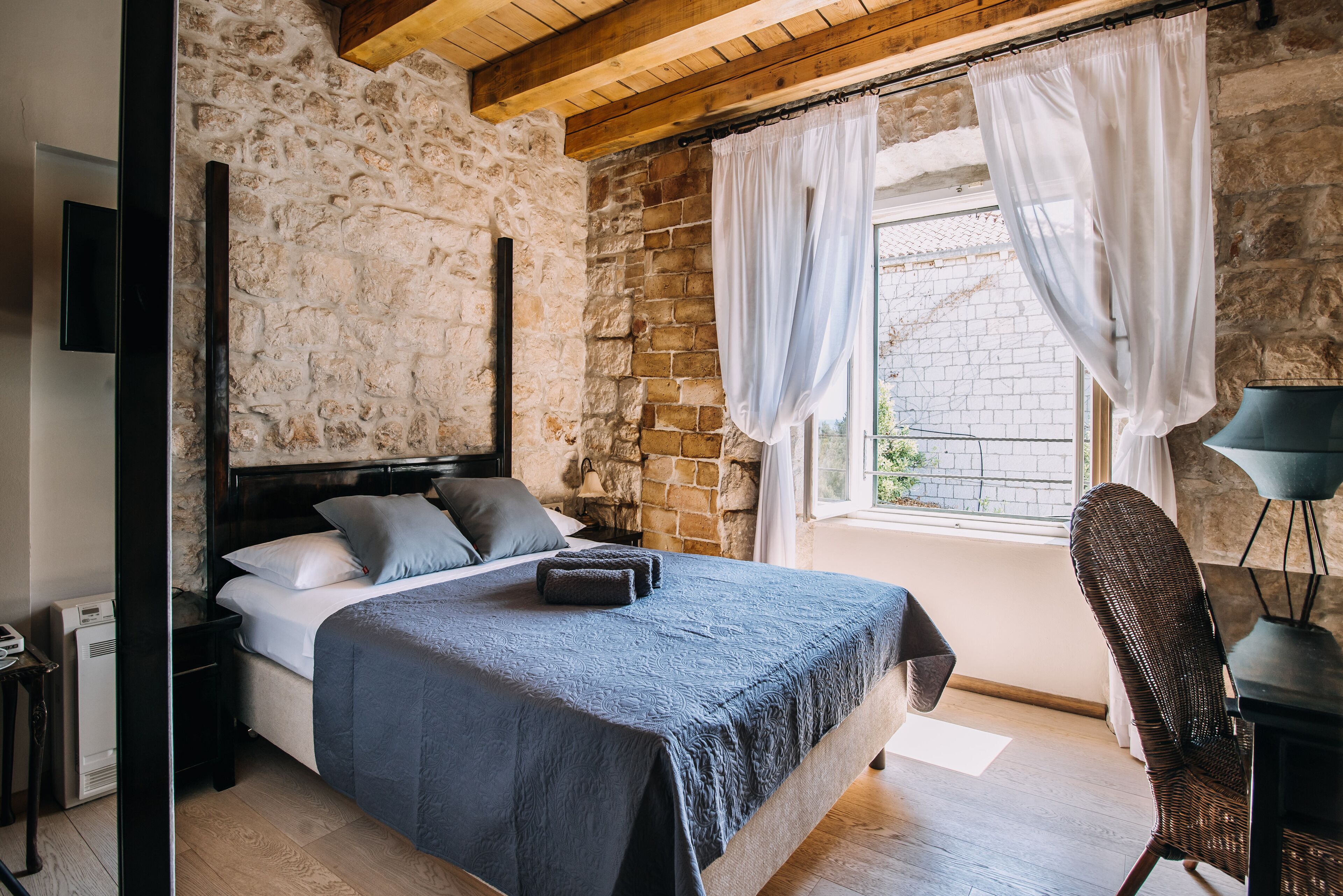 Un dormitorio con pared de piedra, una cama con colcha azul oscuro, una mesita de noche, una silla y una ventana con cortinas.
