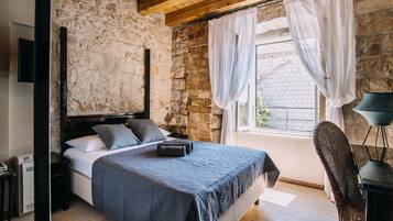 Un dormitorio con pared de piedra, una cama con colcha azul oscuro, una mesita de noche, una silla y una ventana con cortinas.