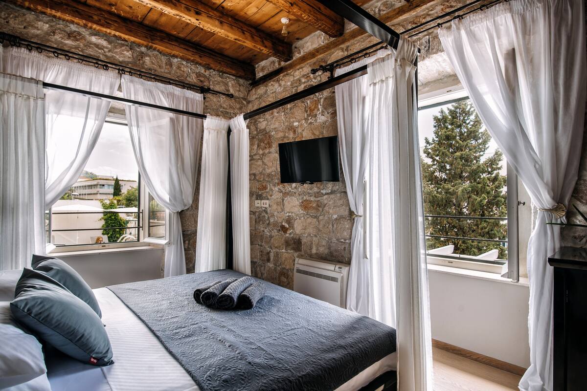 Een slaapkamer met een stenen muur, een bed met een grijze deken, witte gordijnen en een aan de muur bevestigde televisie.