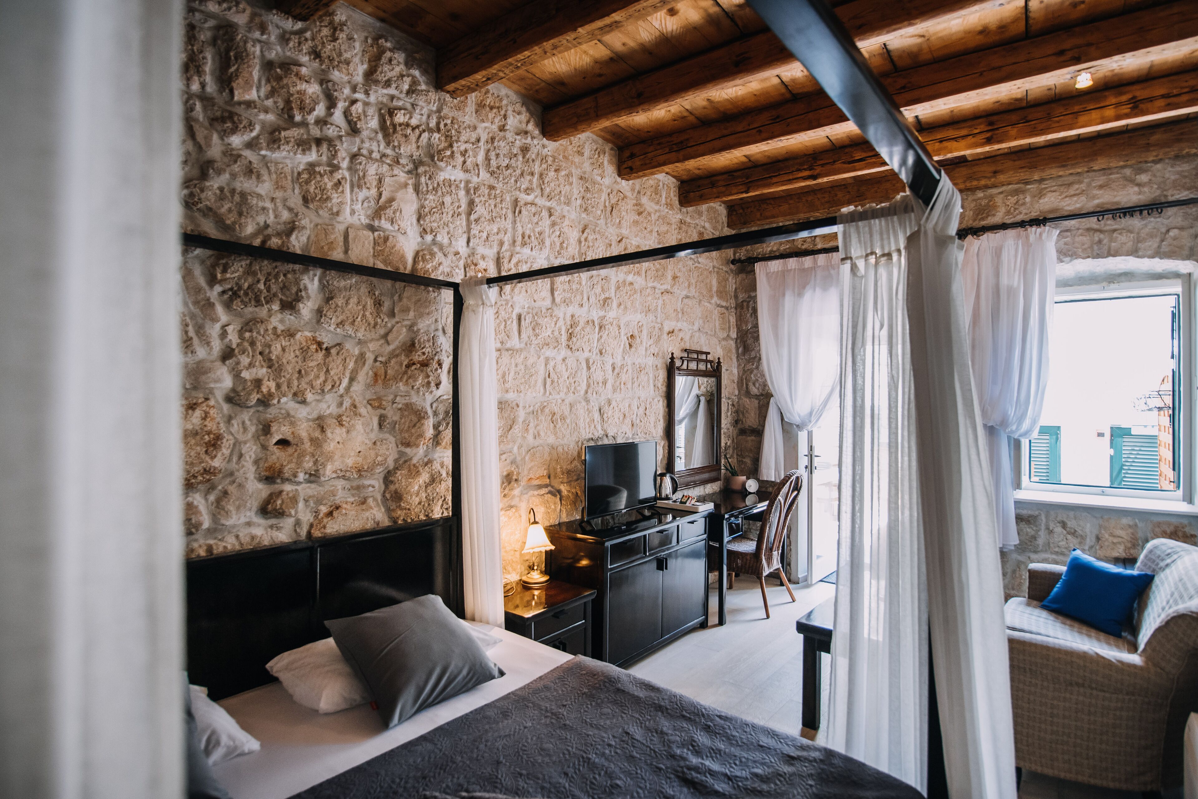 A bedroom with a stone wall, a bed, a desk with a television, and a view of the city through a window.