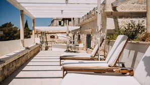 Un espacio al aire libre iluminado por el sol con sillones de descanso y una pérgola, con vista a un edificio de piedra y un cielo despejado.