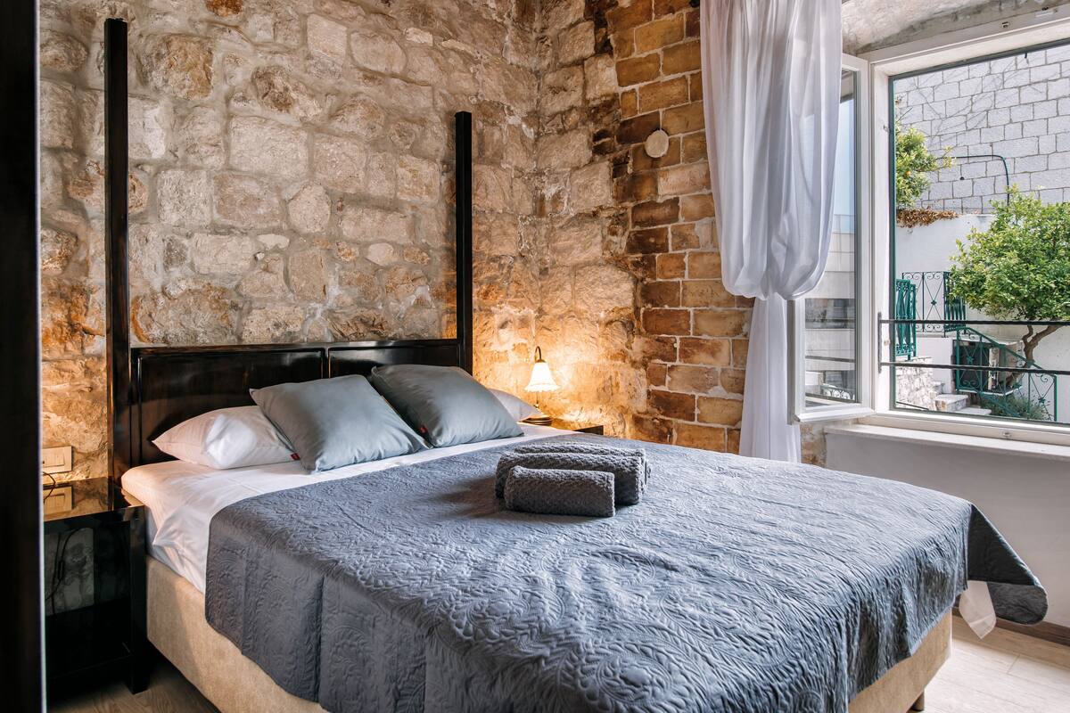 Een slaapkamer met een stenen muur, een bed met grijze beddengoed en een raam met uitzicht op groen.
