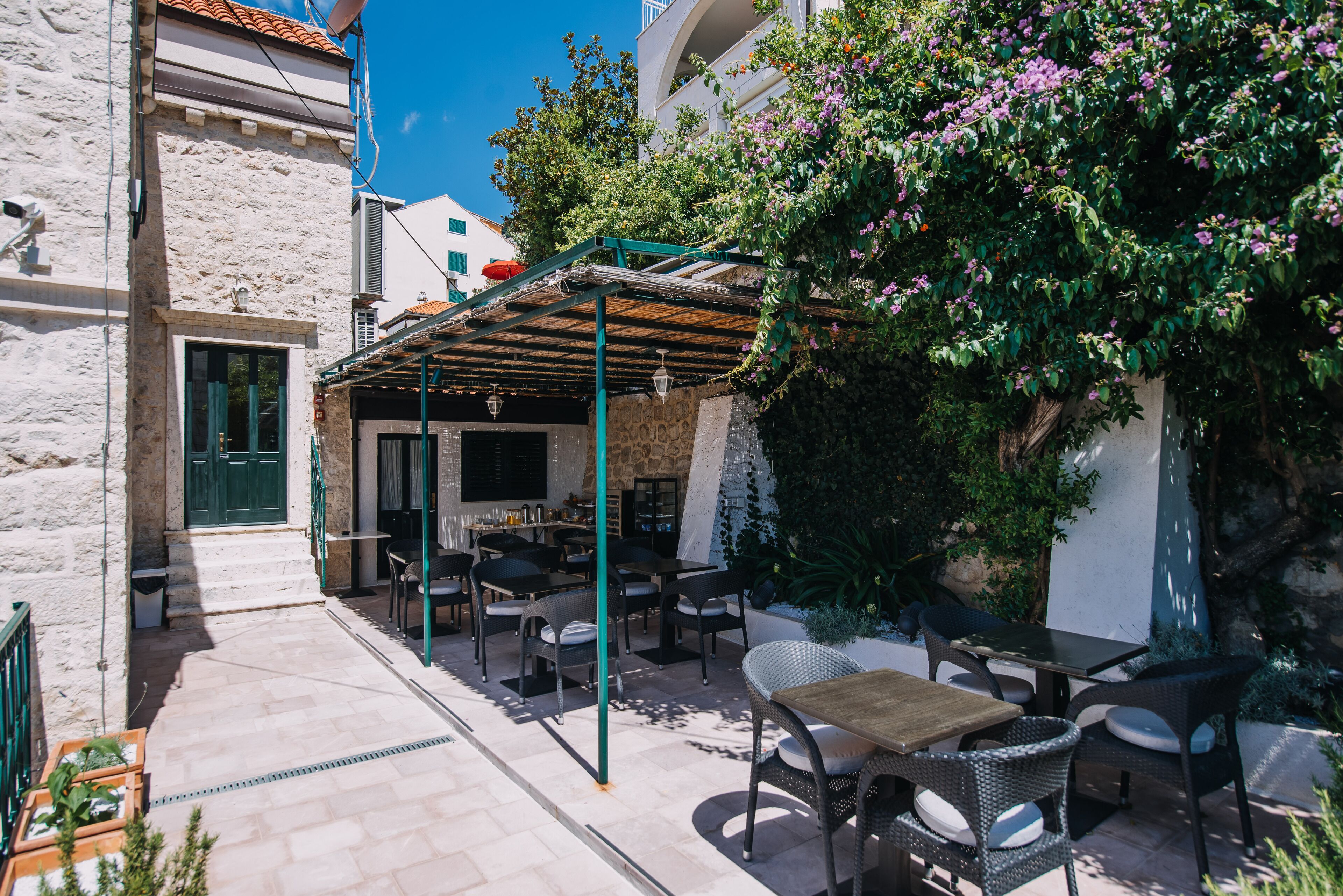 An outdoor cafe with tables and chairs, a stone building, and a covered seating area with a pergola.