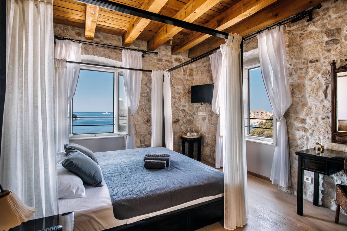 Een slaapkamer met een stenen muur, een bed met grijze beddengoed, een nachtkastje en een televisie aan de muur.