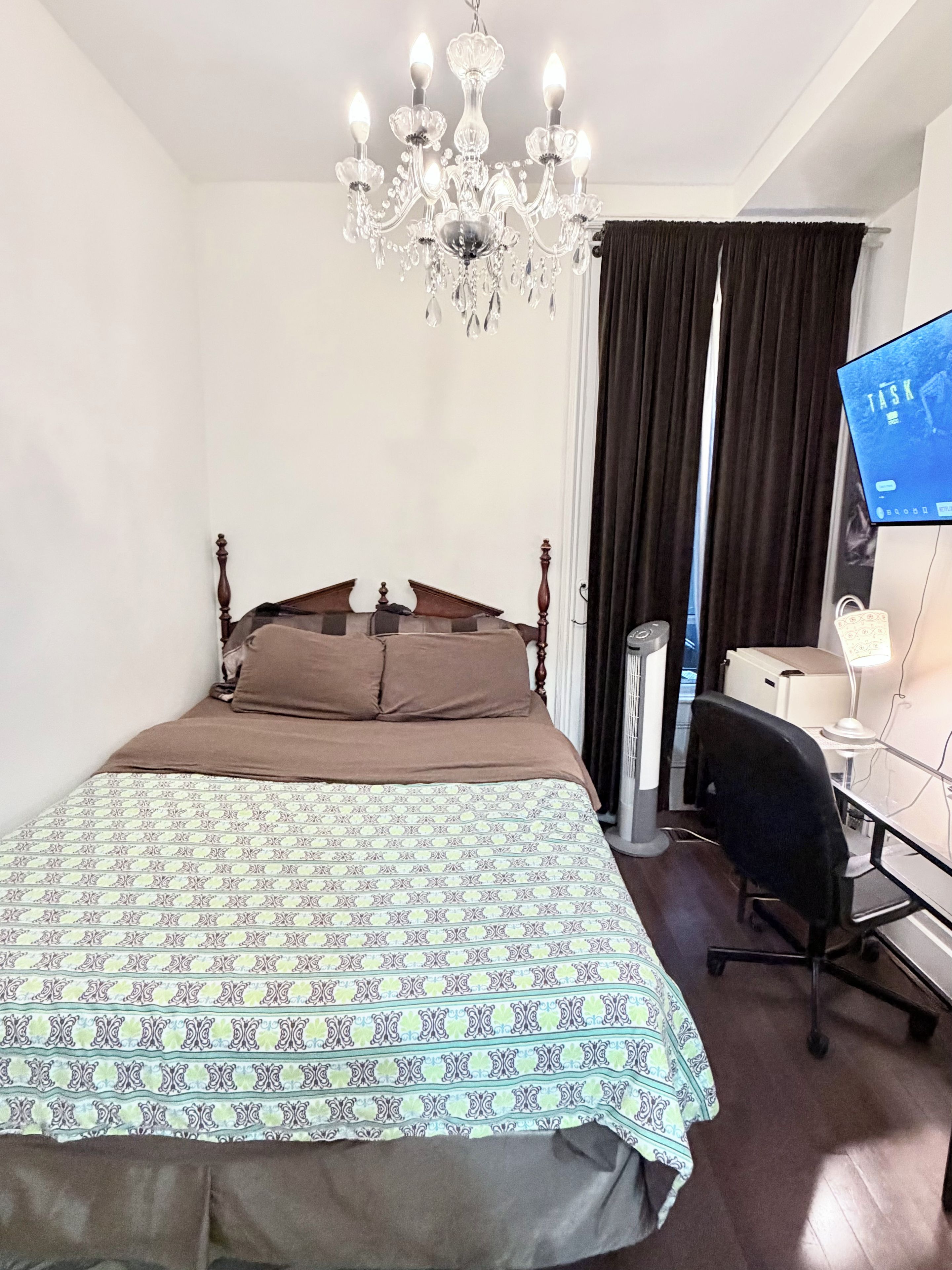 A bedroom with a large bed, a desk, a chair, a computer monitor, and a chandelier.