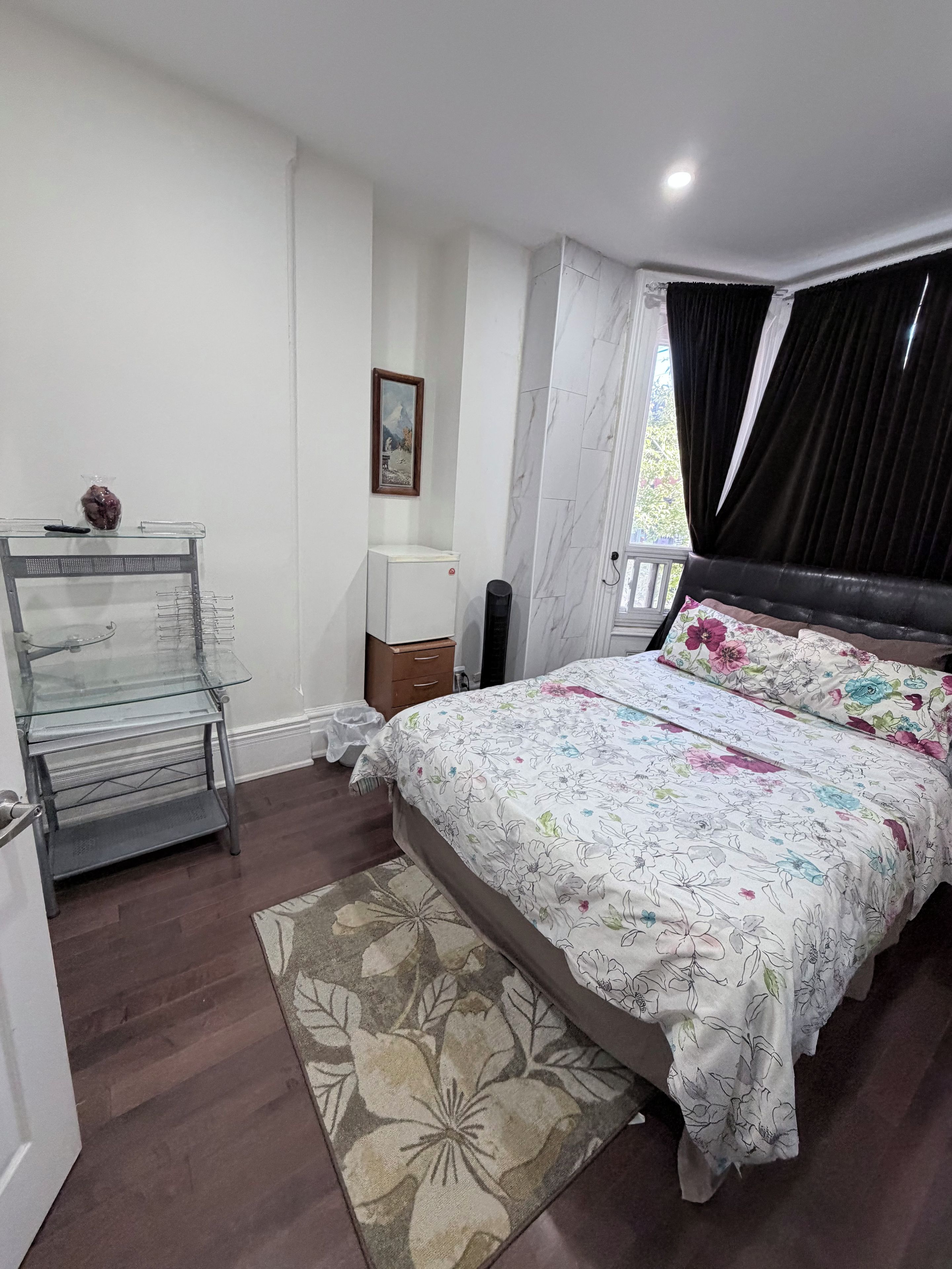 Un dormitorio con cama, mesita de noche, cómoda, vitrina de vidrio, un cuadro en la pared y ventana con cortinas.