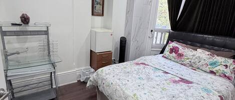 Un dormitorio con cama, mesita de noche, cómoda, vitrina de vidrio, un cuadro en la pared y ventana con cortinas.