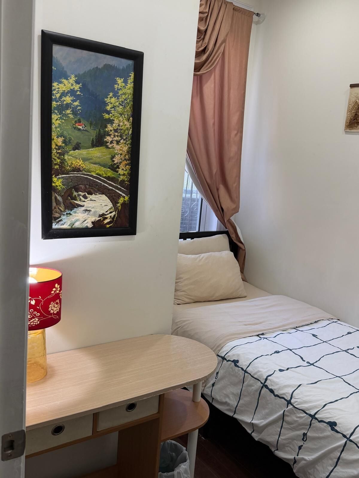 A bedroom with a bed, a desk, a lamp, a window with curtains, and a painting on the wall.