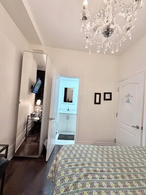 Quarto com cama, lustre, porta com placa e banheiro visível através de uma porta aberta.