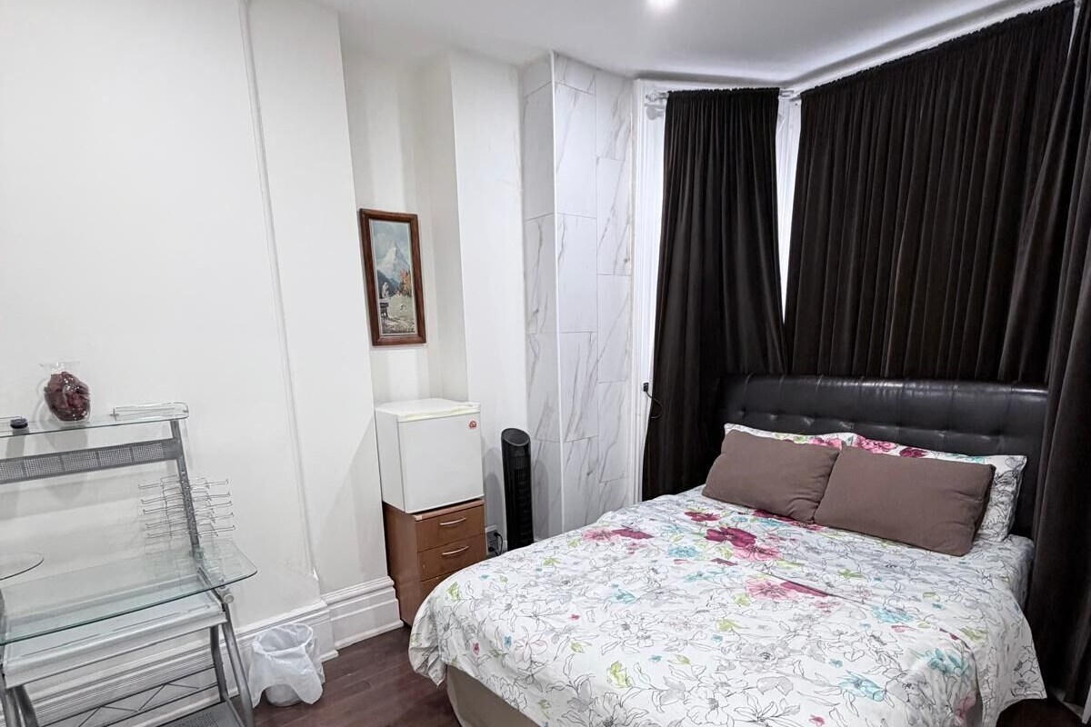 A bedroom with a bed, a desk, a chair, a small table, a lamp, a picture on the wall, and a window with curtains.