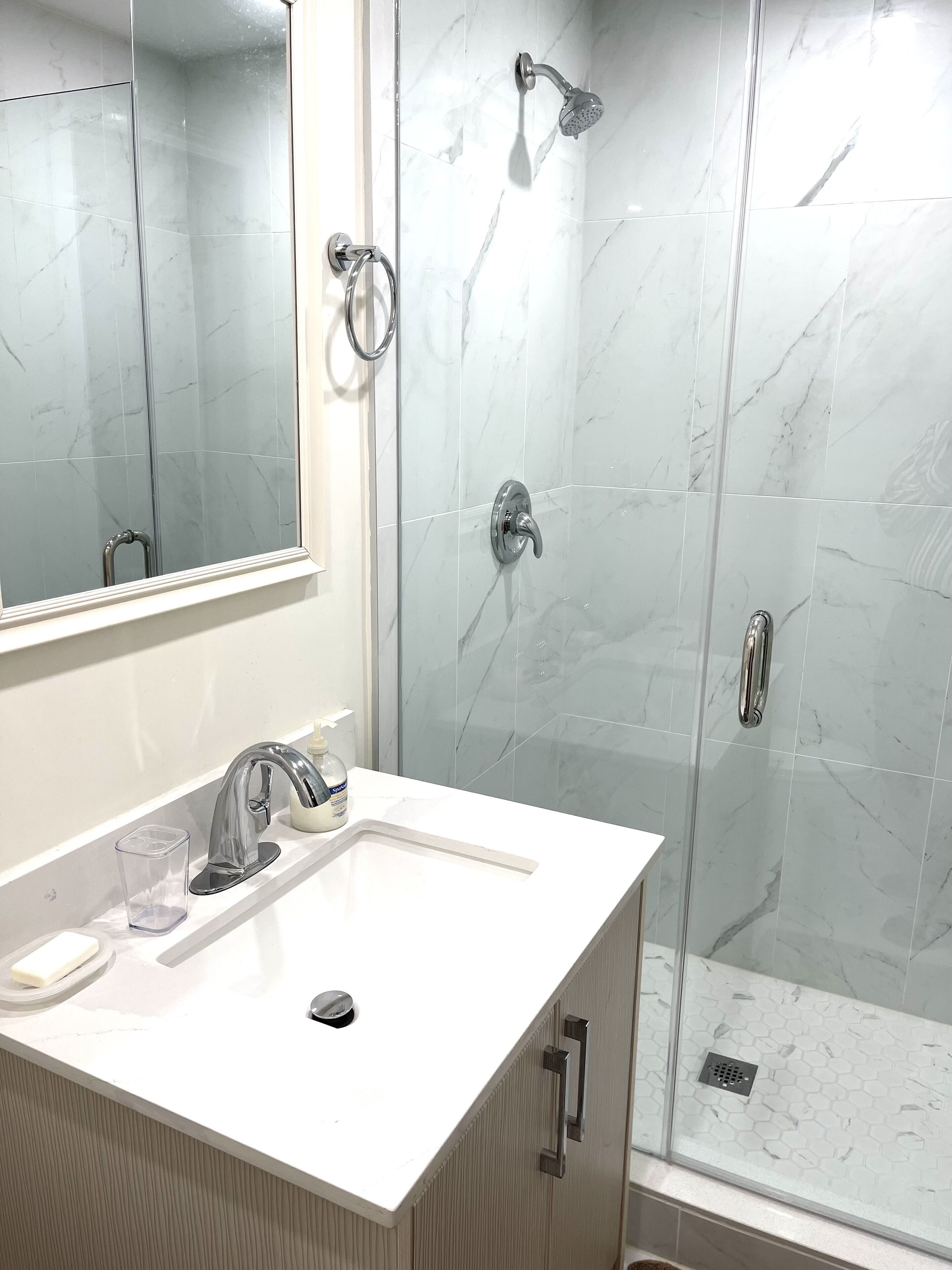 Casa de banho com lavatório branco, espelho e chuveiro com porta de vidro.