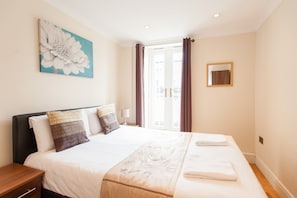 A hotel room with a large bed, a nightstand, a wall-mounted mirror, and a window with curtains. - London Serviced Apartments (London)