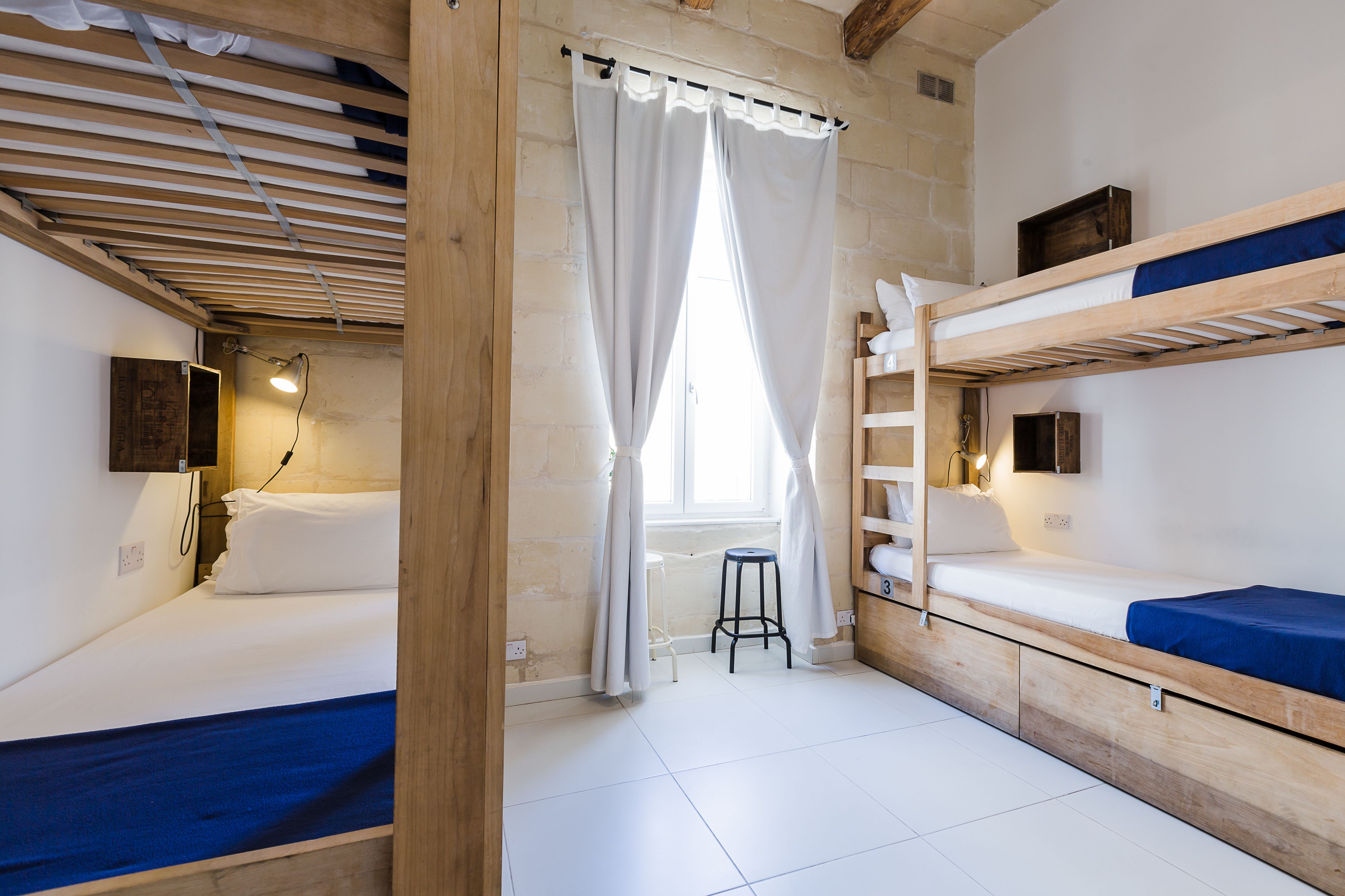 A compact room with two bunk beds, a small stool, a window with white curtains, and a wooden ceiling.