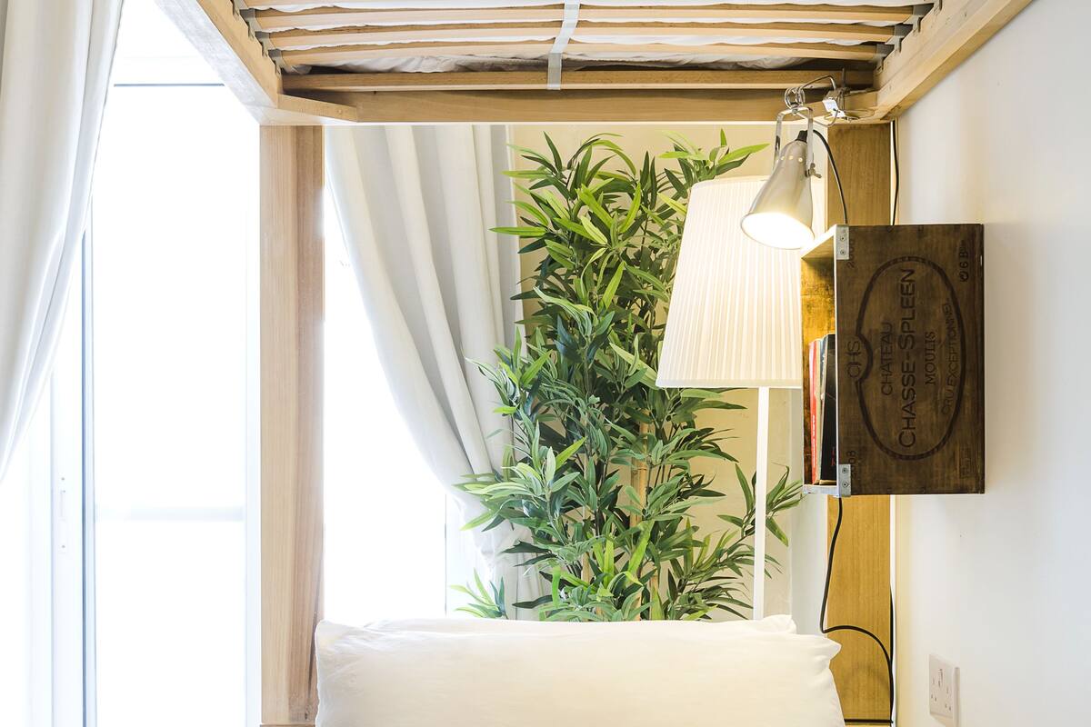 A neatly made bed with a rolled white towel, a potted plant, and a wall-mounted lamp in a room with wooden beams and a window with curtains.