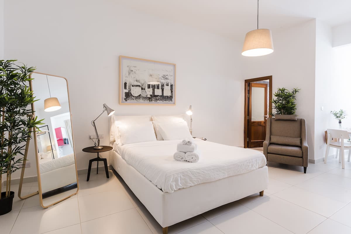 A modern hotel room with a large bed, bedside lamps, a mirror, a potted plant, and a painting on the wall.