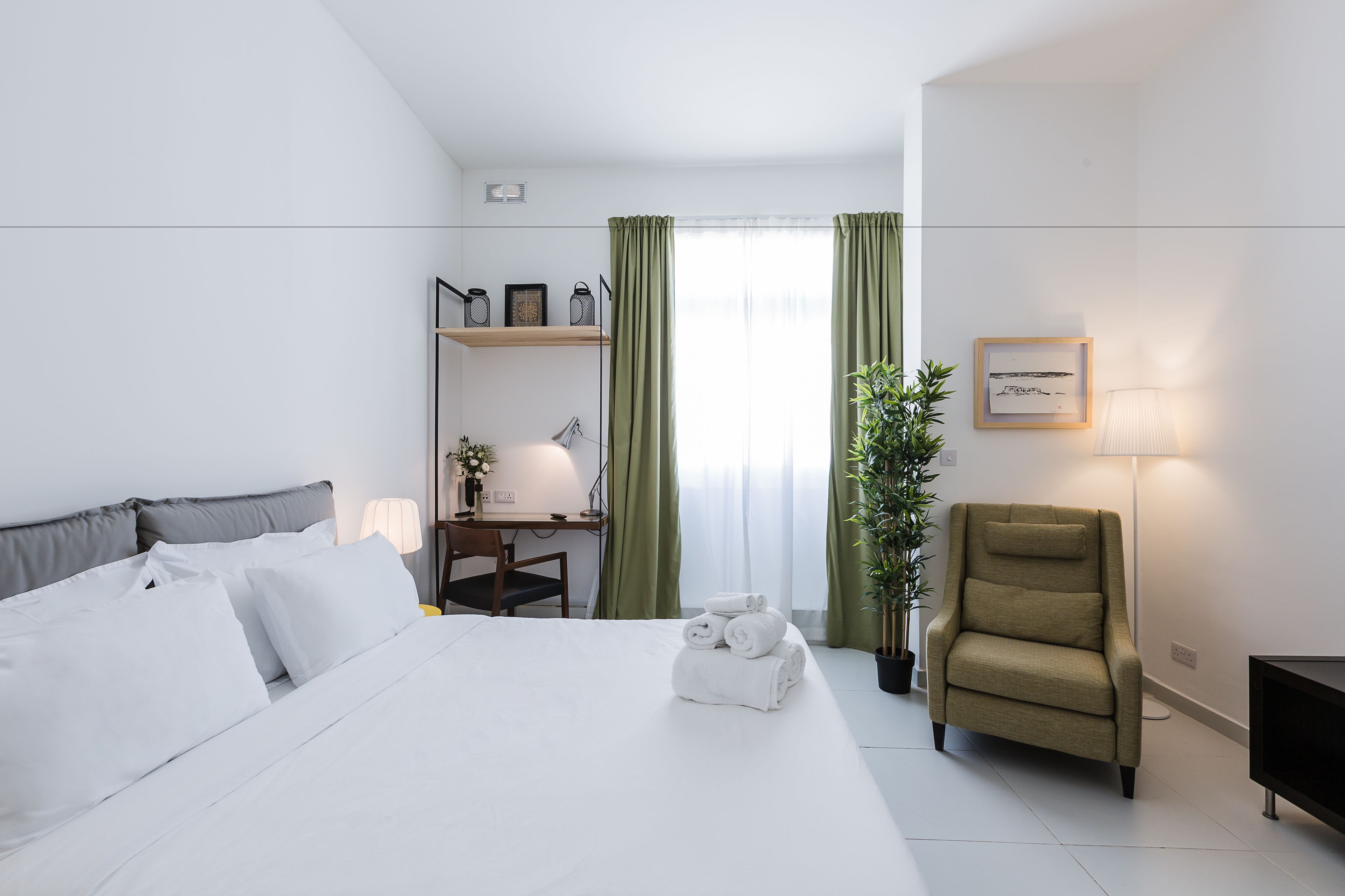 A hotel room with a bed, a desk with a lamp, a chair, and a potted plant.