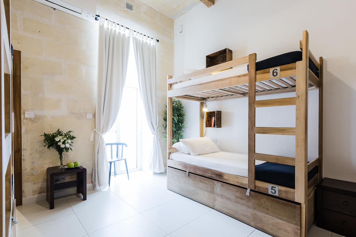 A bunk bed with numbers 5 and 6, a small wooden nightstand, a chair, and a potted plant in a room with white walls and a brick wall.