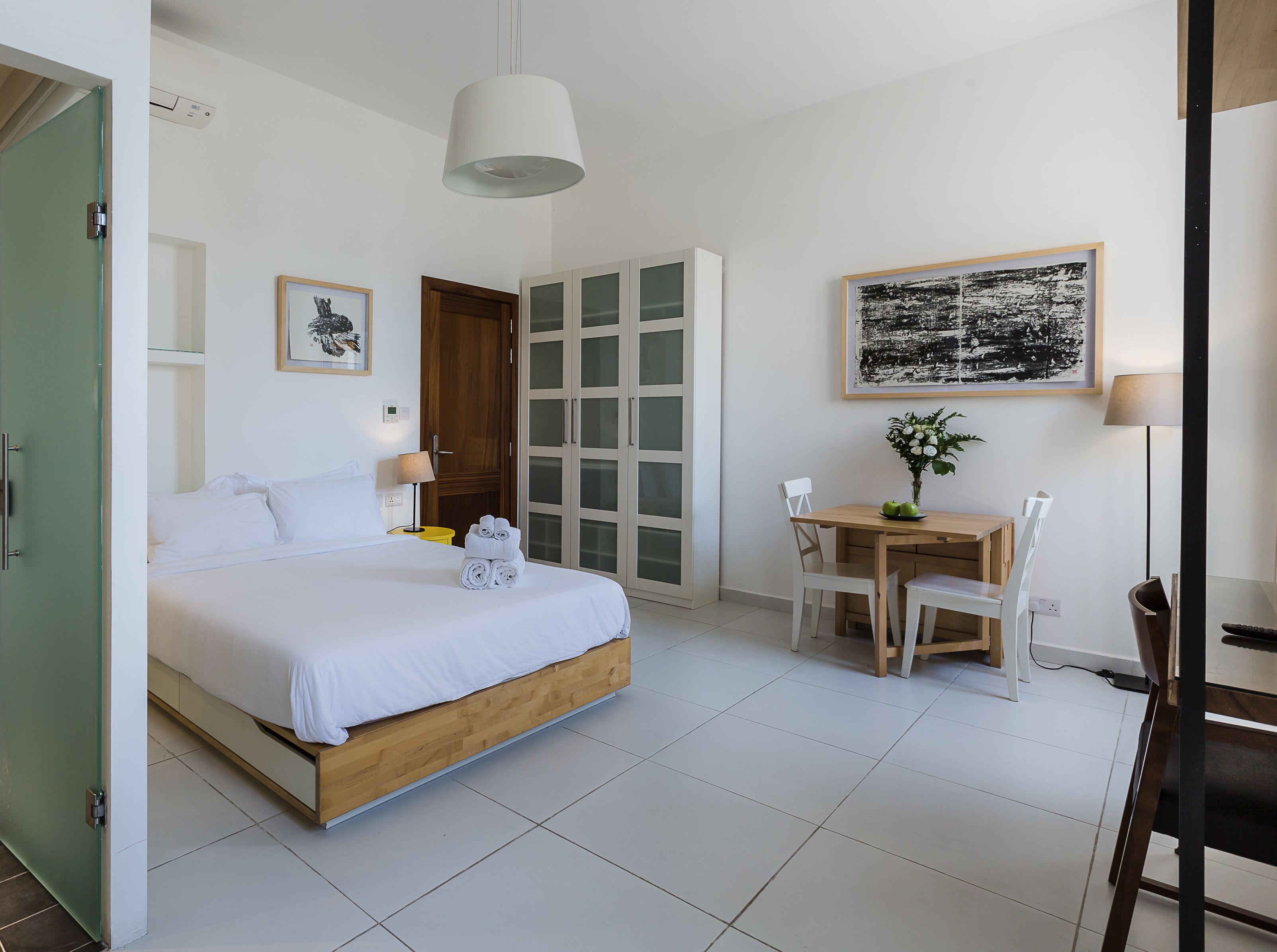 A hotel room with a bed, a desk, and a chair. There is a door, a window, and a painting on the wall.