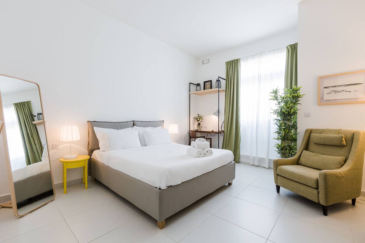 A hotel room with a bed, a chair, a desk, a mirror, and a plant.