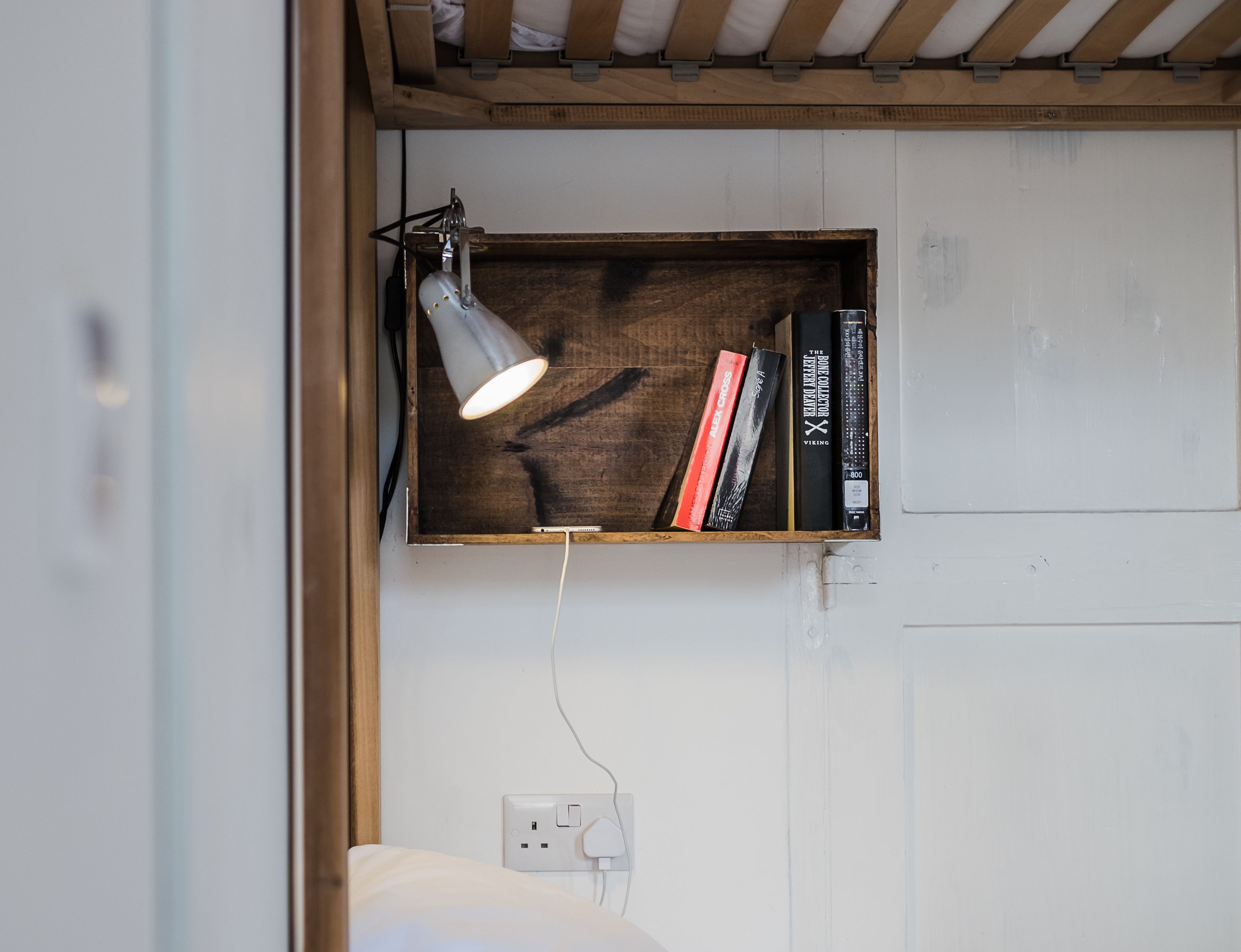 Uno scaffale di legno con libri e una lampada, una presa di corrente e un letto con biancheria bianca.