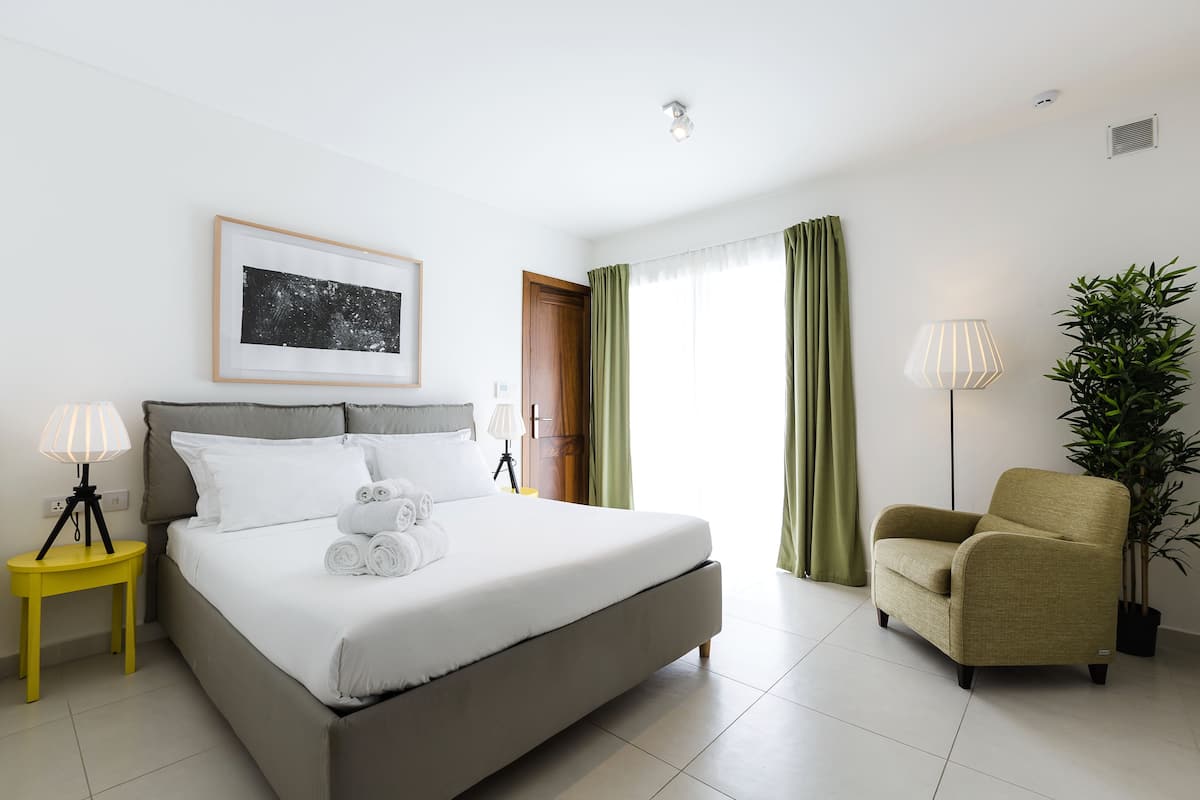 A neatly arranged hotel room with a bed, bedside lamps, a chair, a potted plant, and a framed picture on the wall.