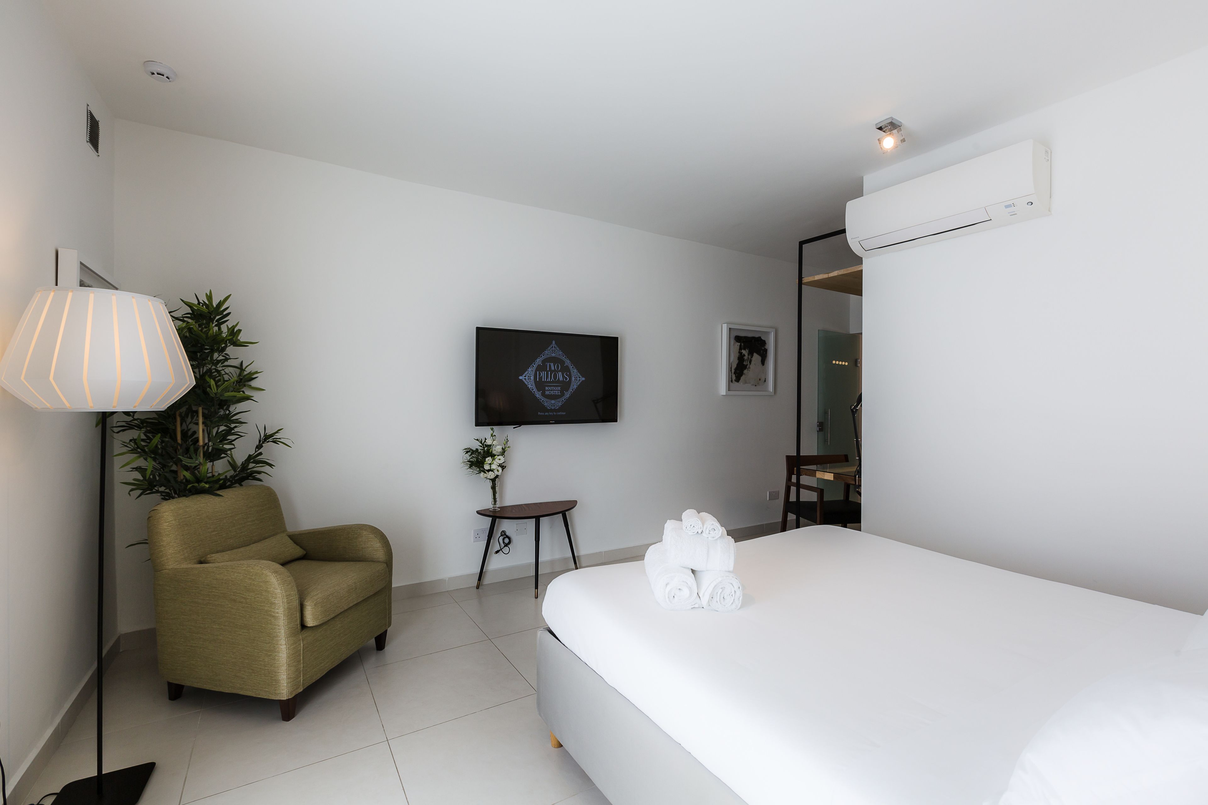 Una camera d'albergo con un letto, una poltrona, un tavolino con una pianta, una TV a parete e un condizionatore d'aria.