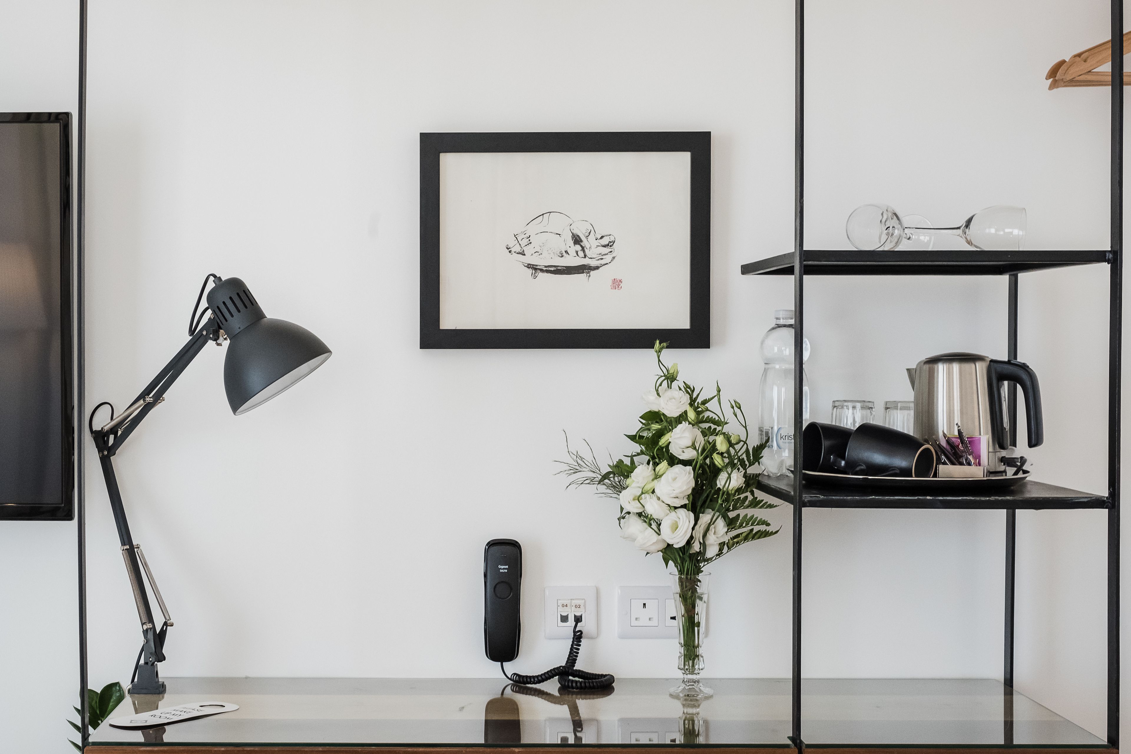 A desk with a lamp, a framed picture, a vase with flowers, and a shelf with kitchen items.