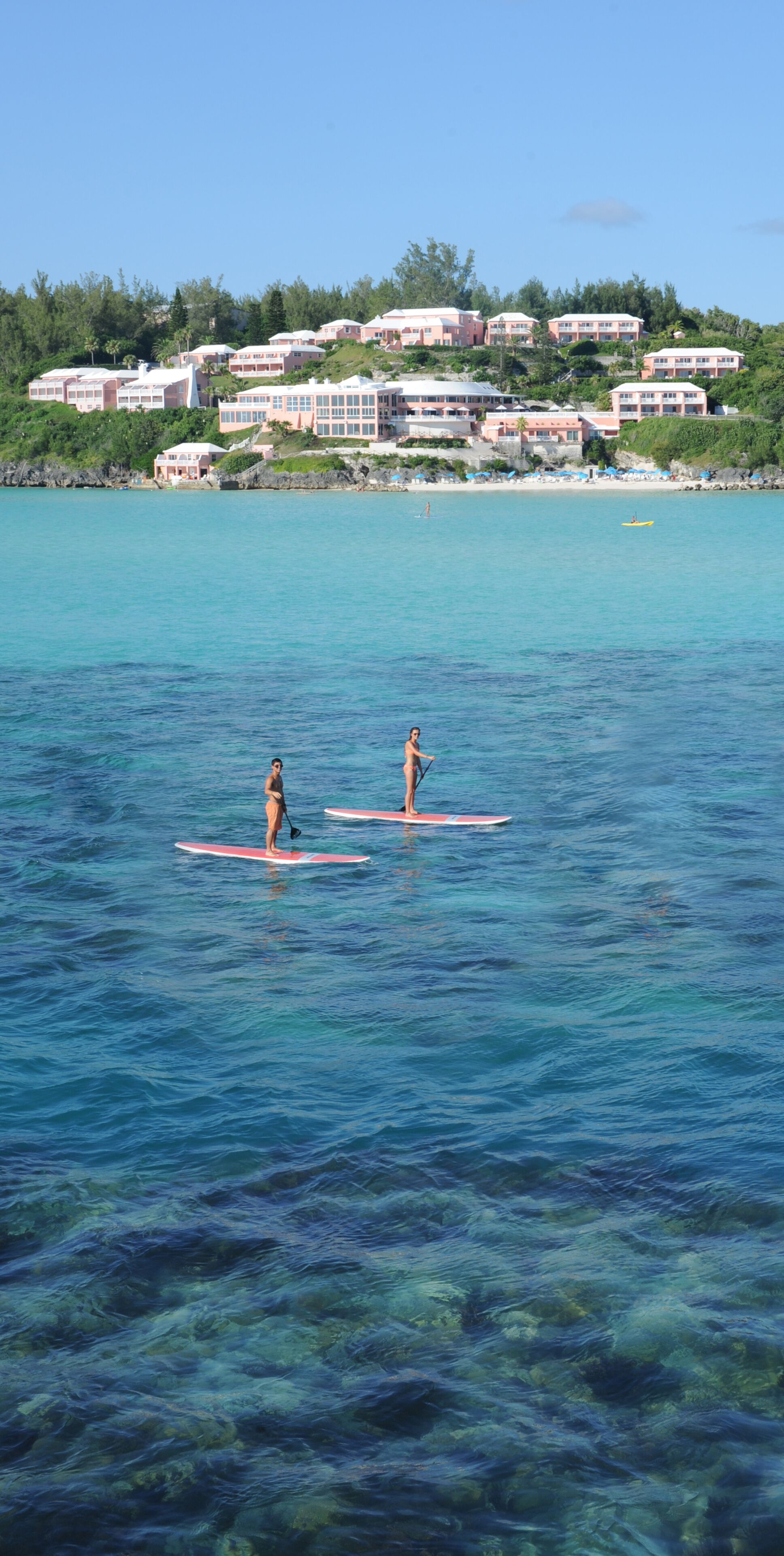 Two people paddleboarding on a clear blue sea with a resort in the background.
