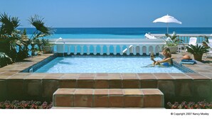 A pool area with a view of the ocean, a white umbrella, and a woman reading a book. - Pompano Beach Club (Southampton Parish)