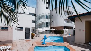 Un área de piscina con sillones de descanso y sombrillas, rodeada de edificios modernos con grandes ventanales y una palmera.