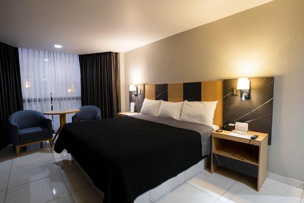 Habitación de hotel con cama, mesita de noche, lámpara, silla y vista al exterior a través de una ventana con cortinas.