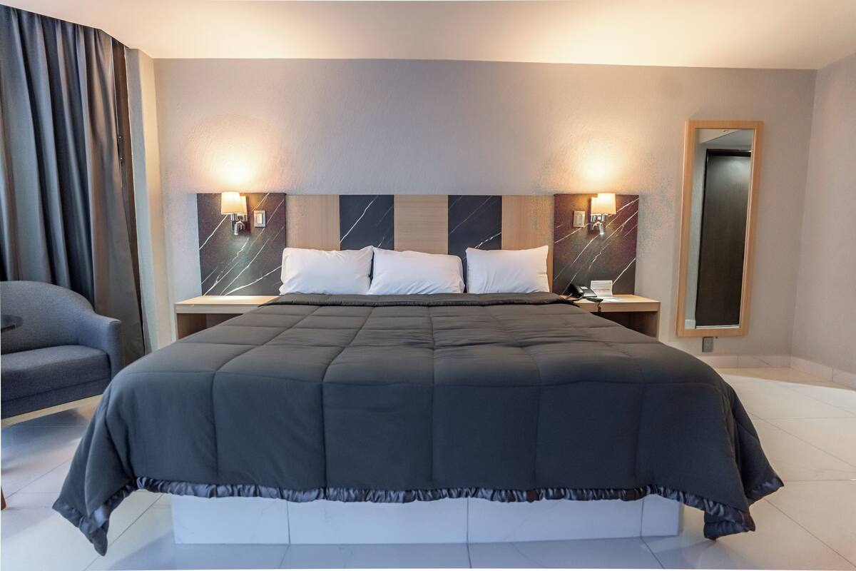 Una habitación de hotel moderna con una cama grande, dos mesitas de noche, una silla y un espejo.