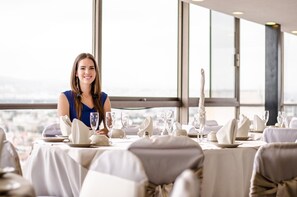 A dining table set with white tableware and glasses, with a city view through large windows.
