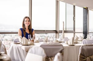 Mesa de comedor dispuesta con vajilla y copas blancas, con vista a la ciudad a través de amplios ventanales.