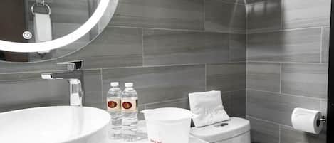 Un baño moderno con un lavamanos grande, dos botellas de agua, un cubo blanco, un inodoro y un perchero para toallas.