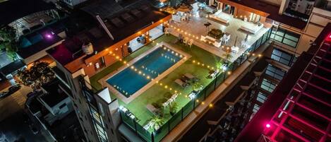 Un área de piscina en la azotea con múltiples sillones de descanso y sombrillas, rodeada de edificios.