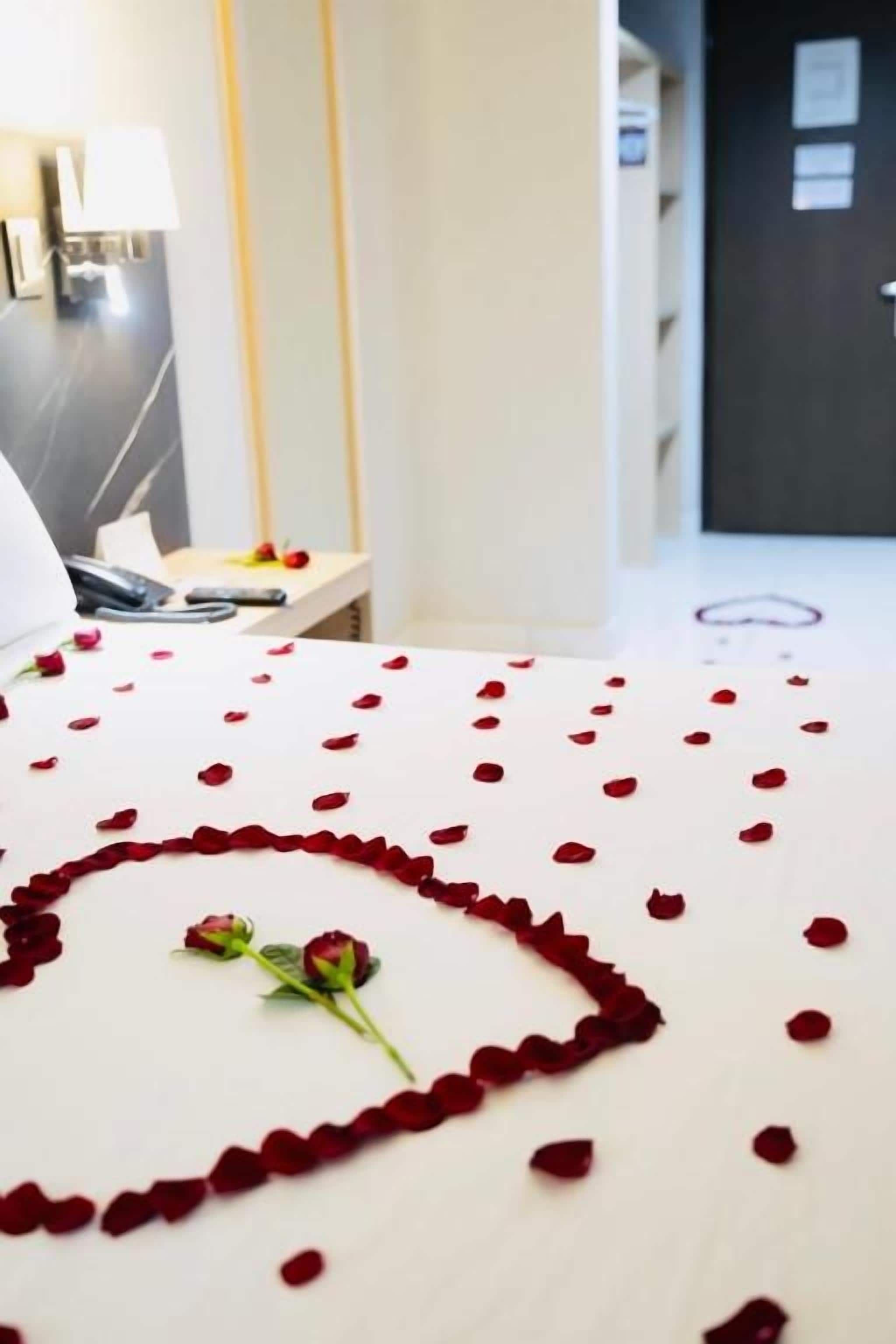 Una habitación de hotel con una cama cubierta de pétalos de rosa y una sola rosa, una mesita de noche con un teléfono y una lámpara de pared.