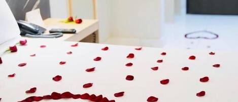 Una habitación de hotel con una cama cubierta de pétalos de rosa y una sola rosa, una mesita de noche con un teléfono y una lámpara de pared.