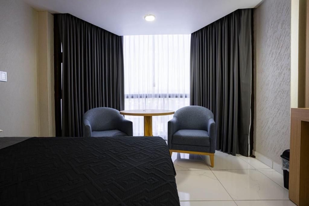 Ett hotellrum med en säng, två fåtöljer, ett litet runt bord och ett fönster med gardiner.