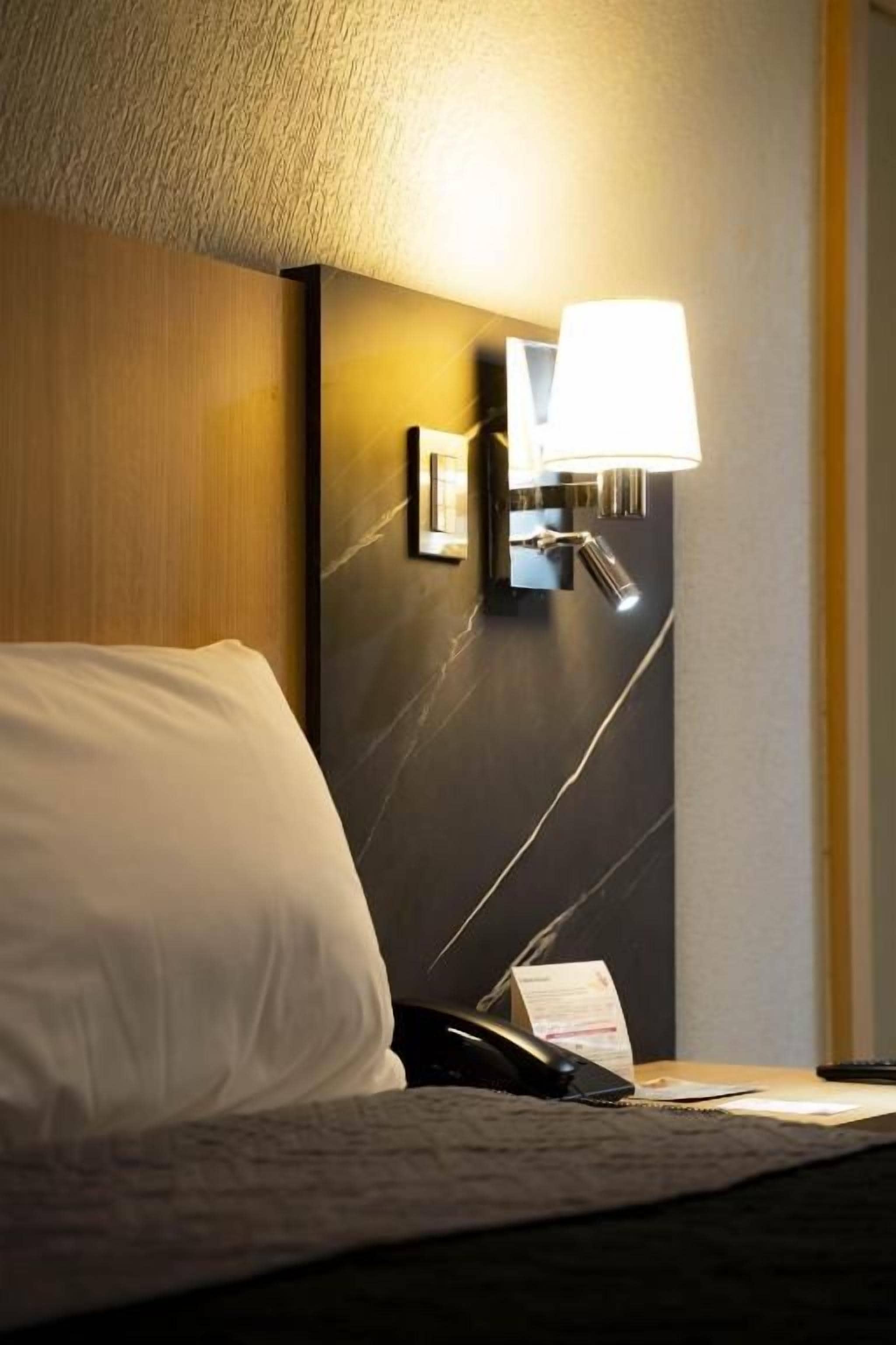 Quarto de hotel com cama, luminária de mesa, telefone e arandela.