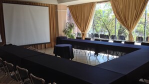 Un salón de conferencias con una larga mesa, sillas, una pantalla de proyector y ventanas grandes con cortinas.