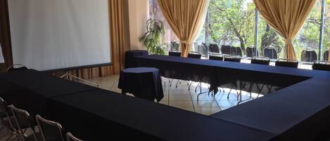 A conference room with a long table, chairs, a projector screen, and large windows with curtains.