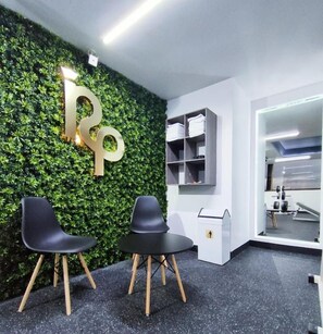 A reception area with a green wall, two black chairs, a white desk, and a mirror reflecting gym equipment.
