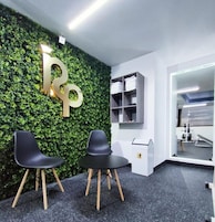 Un área de recepción con una pared verde, dos sillas negras, un escritorio blanco y un espejo que refleja equipo de gimnasio.