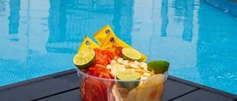 A poolside setting with a clear view of the pool, a table with snacks, and two people toasting with drinks.