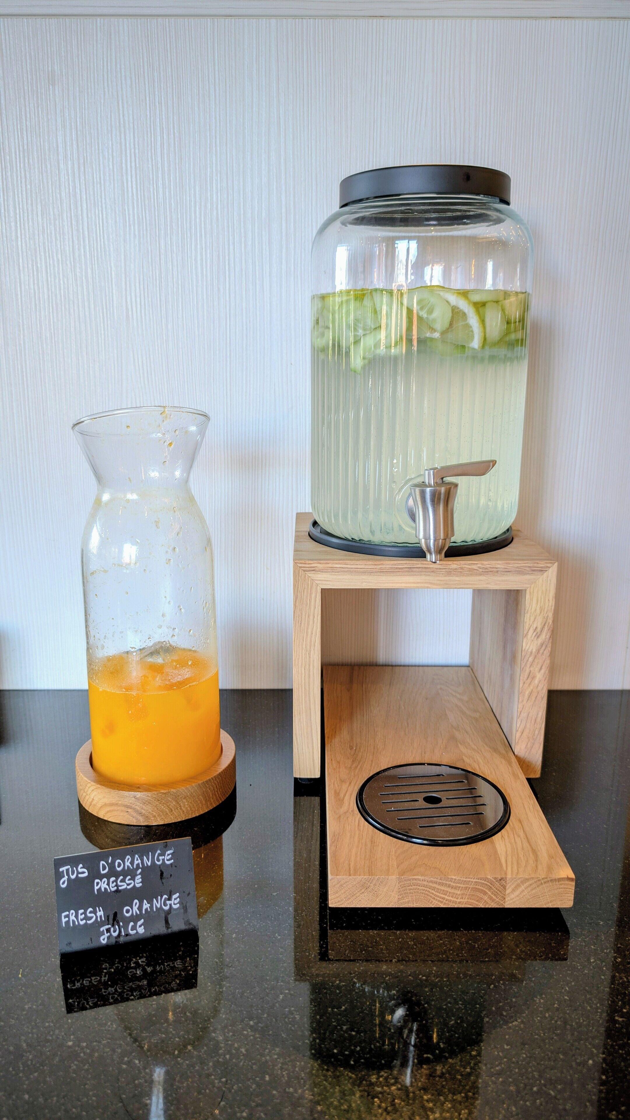 A juice dispenser with a wooden base and a metal spout, and a glass carafe with a metal lid and a label that reads "Jus d'orange pressé" (fresh orange juice).