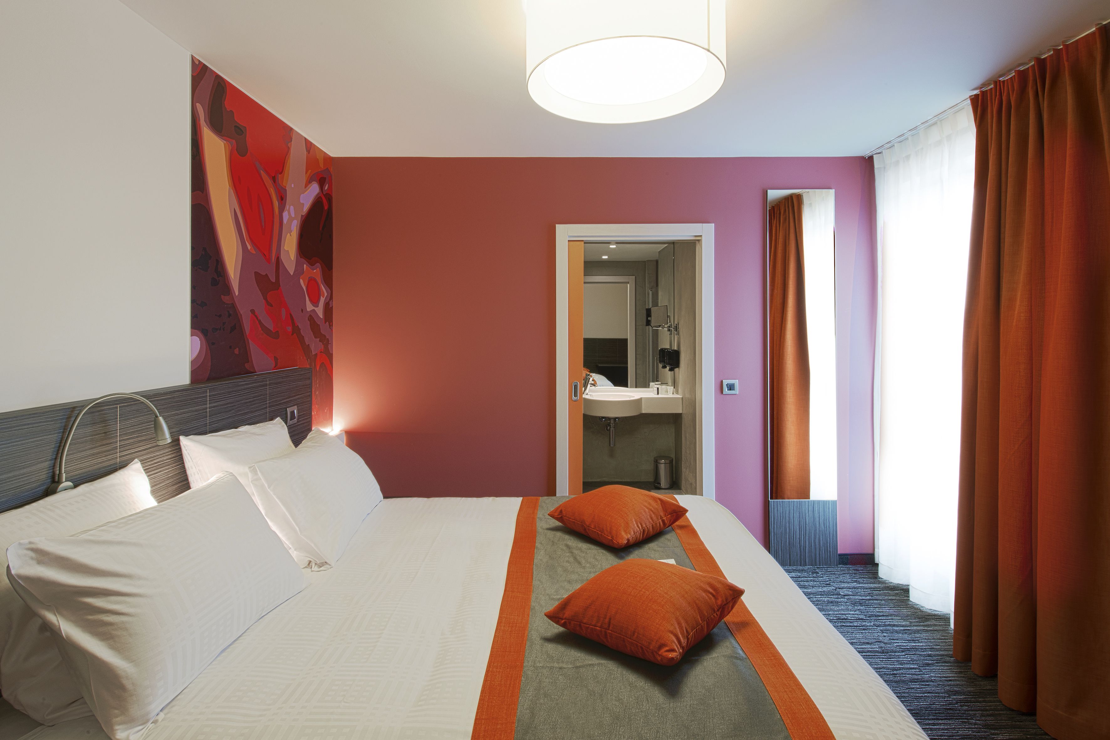 Habitación de hotel con cama, almohadas, mesita de noche, espejo, baño con lavamanos y ventana con cortinas.